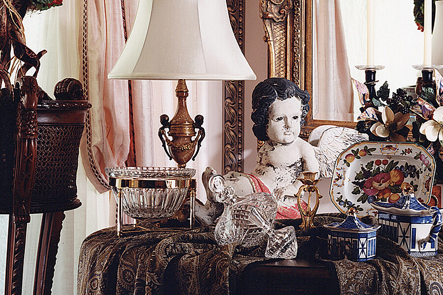 a corner table topped with antiques sits in front of a mirror and draped windows.